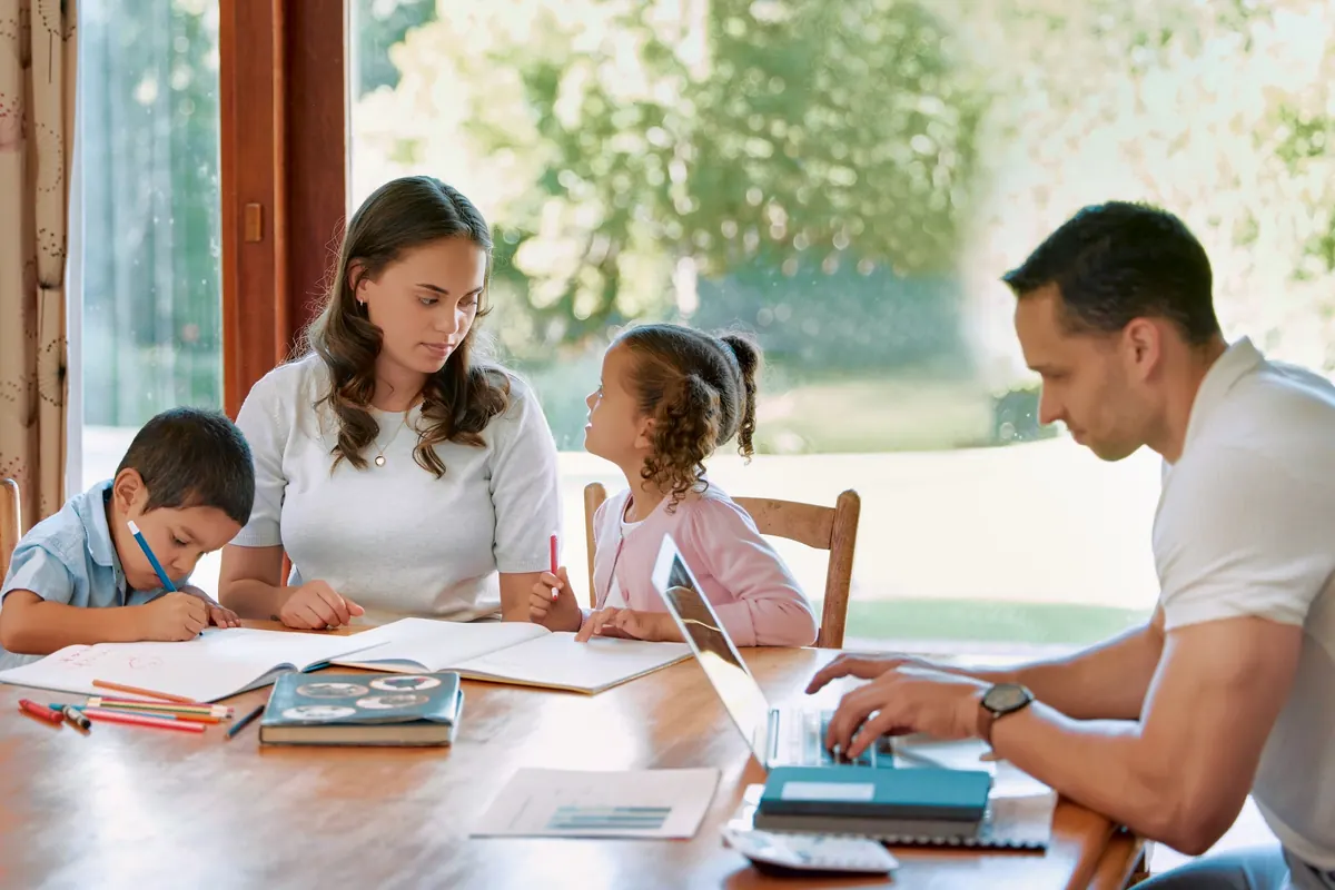 A family at a dining table: two children drawing next to their mother while their father works on a laptop