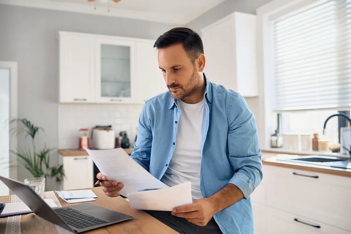 A man in his kitchen reading a letter, with a laptop open on the table
