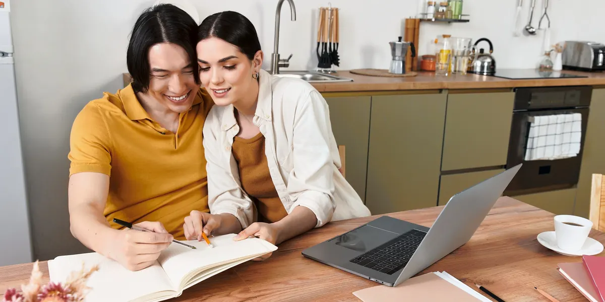 A couple at a kitchen table writing in a notebook, with a laptop open beside them